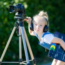 A little girl in headphones with camera on a tripod is standing in a city park.